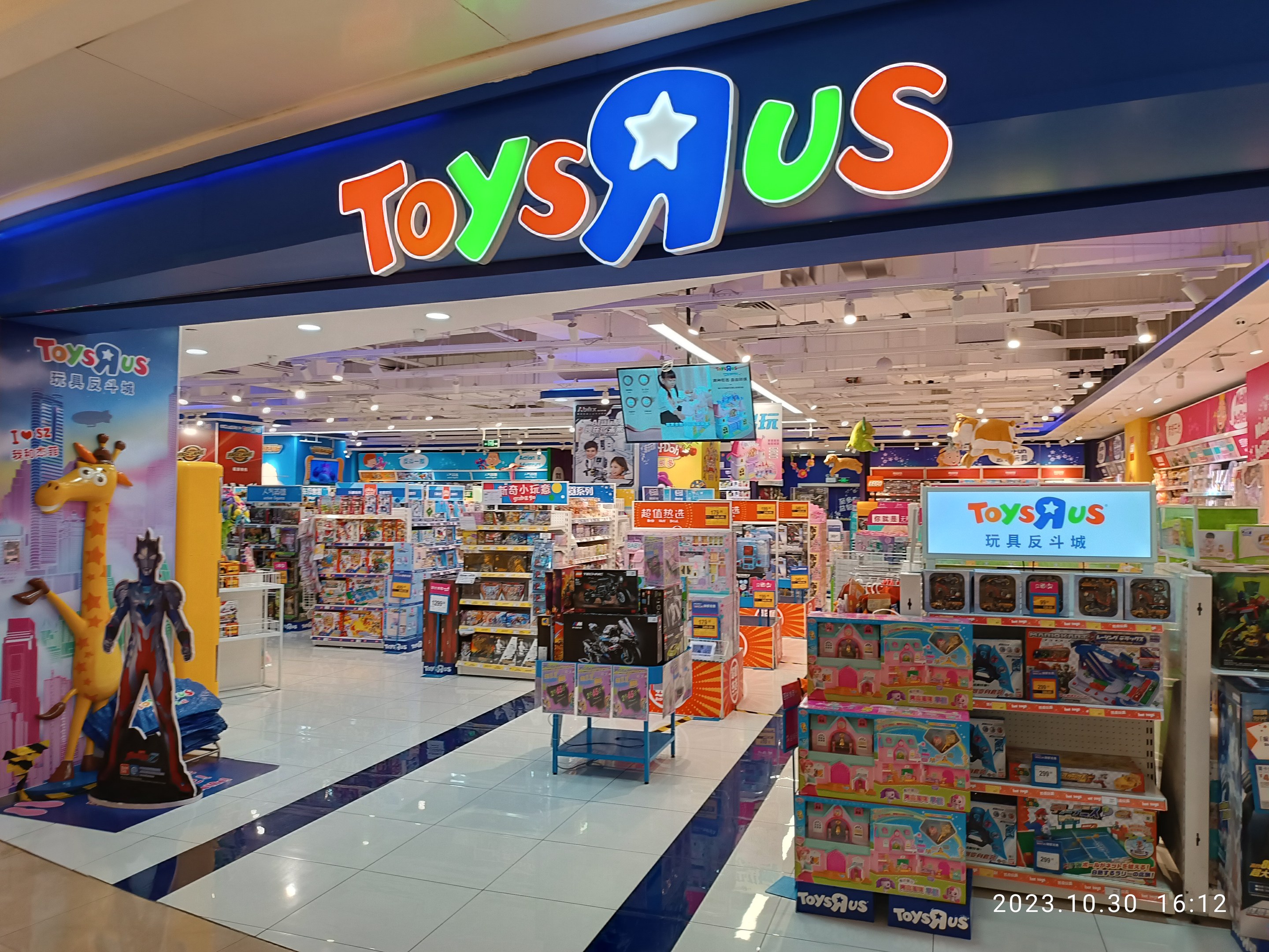 Ein Toys R Us-Laden in einem Einkaufszentrum, der Regale mit Spielzeug und Gegenständen zeigt, ein sichtbares Ladenschild, eine Deckenbeleuchtung und ein Wasserzeichen in der unteren rechten Ecke.