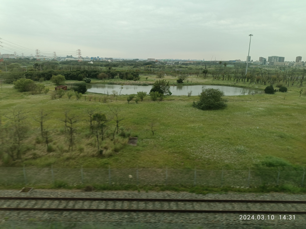 Blick auf einen See aus einem Zugfenster, mit einer Bahnstrecke im Vordergrund, umgeben von Grün, einem Zaun, fernen Gebäuden, Strommasten und einem klaren Himmel.