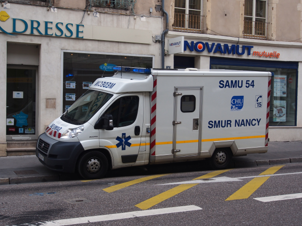 Ambulanz auf dem Seitenstreifen vor einem Gebäude mit Krankenhausschild und verschiedenen Aushängen geparkt.