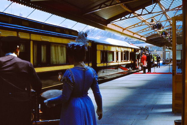 Frau in blauem Kleid neben einem Zug an einem Bahnhof mit einigen Menschen auf dem Bahnsteig.