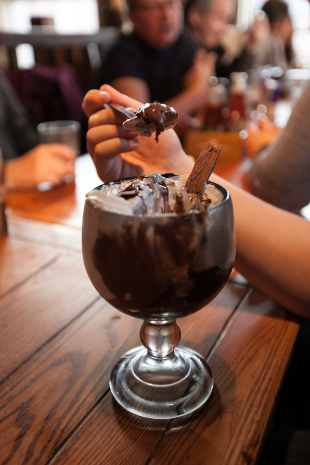 Eine Gruppe von Menschen sitzt um einen Tisch mit einem Glas Schokoladenpudding in der Mitte; eine Person im Vordergrund hält einen Löffel und isst den Pudding, während der Hintergrund unscharf ist.