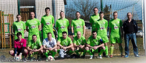 Eine Gruppe von Männern in grünen T-Shirts steht auf einem Fußballfeld mit einem Ball vor ihnen, einige in Hockstellung, in der Nähe eines Torpfostens, mit Bäumen, Gebäuden und Fahrzeugen im Hintergrund.