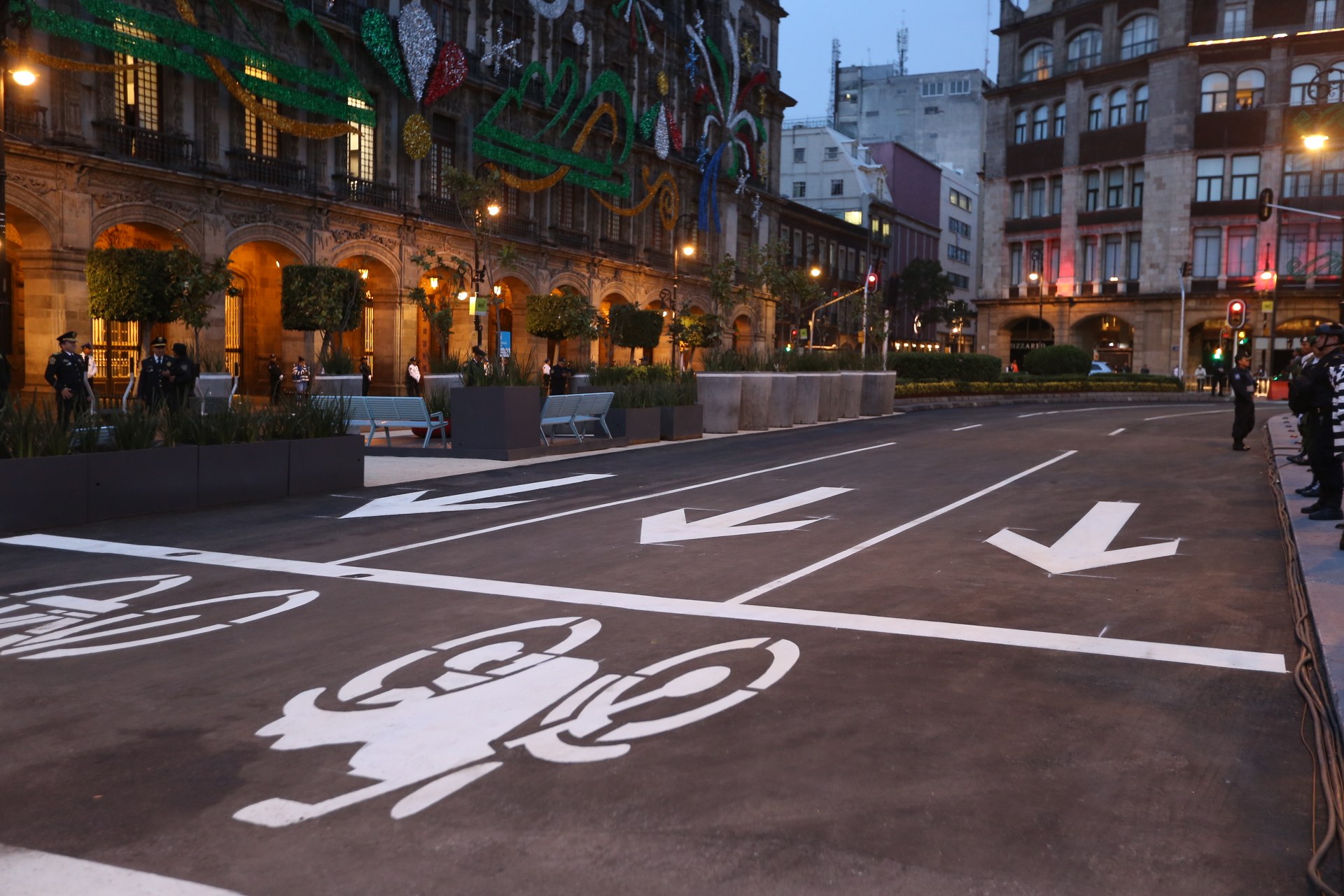 Stadtstraße mit einer ausgewiesenen Fahrradspur, umgeben von Gebäuden und Straßenmöblierung, Menschen auf dem Gehweg und einem klaren blauen Himmel.