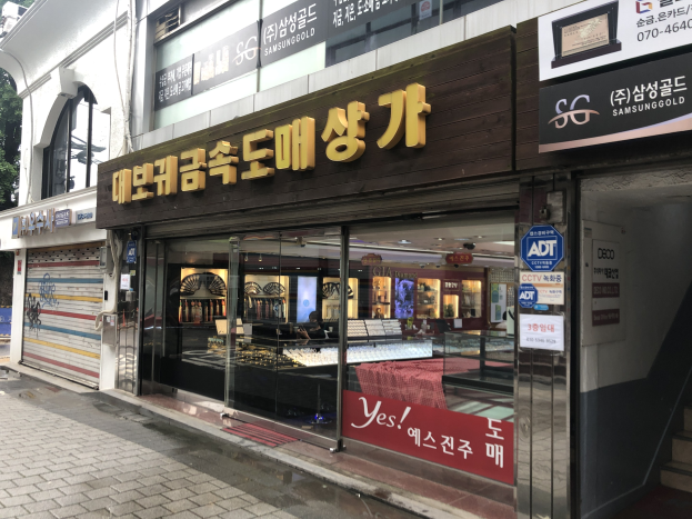 Ladenfront mit einem Schild an der Seite, umgeben von Gebäuden, Bäumen und einer Straße, das verschiedene Gegenstände im Inneren zeigt.