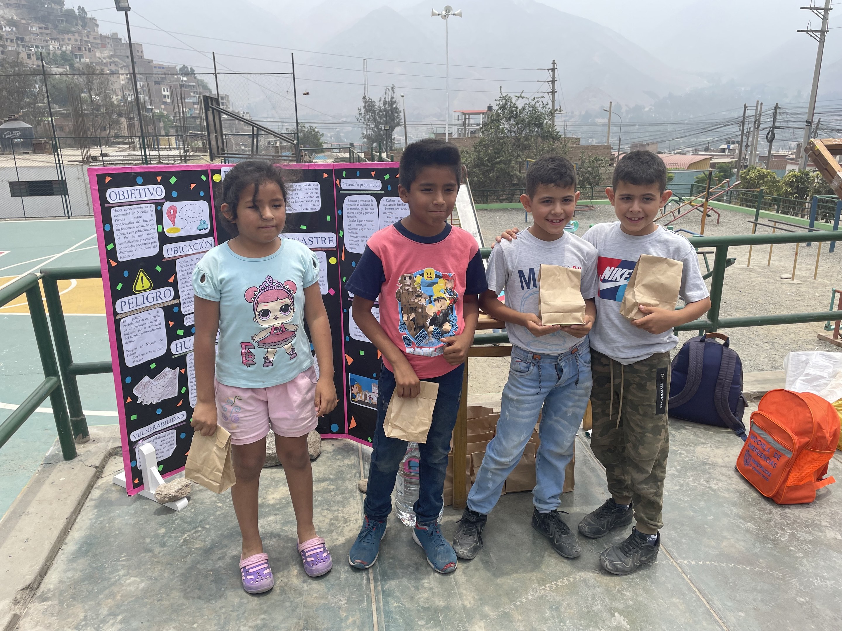 Gruppe von Kindern auf einem Basketballfeld, die Papiertüten halten, mit Tüten und Gegenständen rechts und einer Tafel mit Text links, vor einem Hintergrund aus Zäunen, Pfosten, Straßenlaternen, Bäumen, Gebäuden, Hügeln und Himmel.