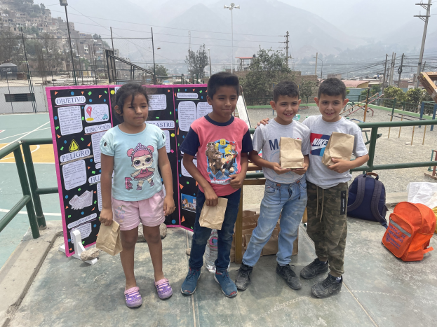 Gruppe von Kindern auf einem Basketballfeld, die Papiertüten halten, mit Tüten und Gegenständen rechts und einer Tafel mit Text links, vor einem Hintergrund aus Zäunen, Pfosten, Straßenlaternen, Bäumen, Gebäuden, Hügeln und Himmel.