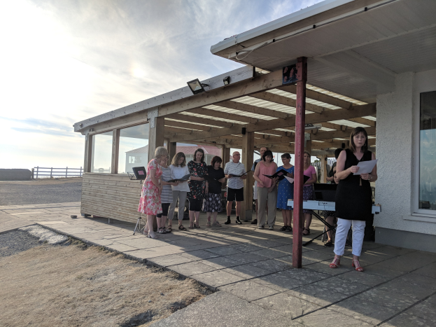 Eine Gruppe von Menschen versammelt sich vor einem modernen Gebäude mit Glasfenstern und Säulen, einige halten Papiere, mit einem Zaun und bewölktem Himmel im Hintergrund, wahrscheinlich feiern sie die Eröffnung eines neuen Gemeindezentrums.