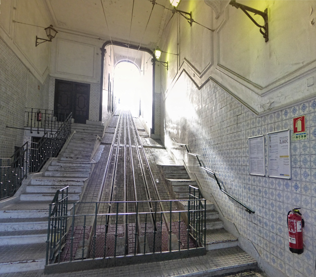 Alte Bahnstation mit Treppe, die zu einer Gebäudeterrasse führt, mit Geländern, Wandtafeln, Feuerlöscher, Deckenlampen und einer Tür im Hintergrund.