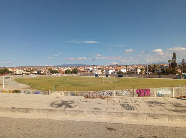 Fußballfeld mit Graffiti an der Seite, umgeben von einer Straße, Gras, Pfählen, Bäumen, Gebäuden und einem bewölkten Himmel.
