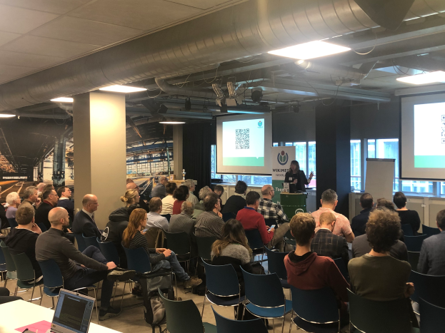 Eine große Gruppe von Menschen, die auf Stühlen sitzen und auf eine Projektionsfläche schauen, mit einem Redner an einem Podium mit Mikrofon und Gerät in einem Raum mit einer Tafel, Säulen, Fenstern, Deckenleuchten und Dach, an einer Workshop-Veranstaltung über Blockchain-Technologie teilnehmen.