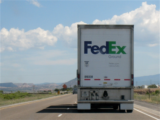 FedEx-Lkw auf einer Highway mit Bergen im Hintergrund, umgeben von Bäumen, Pflanzen, Pfählen und Brettern, mit Text unten.