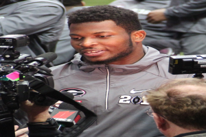 Ein Mann in einem grauen Jackett hält eine Videokamera und spricht mit Reportern, während mehrere Personen im Hintergrund zu sehen sind.