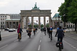 Eine Gruppe von Menschen, die Fahrräder vor dem Brandenburger Tor in Berlin, Deutschland, fahren, mit Bäumen, Laternenmasten und Gebäuden im Hintergrund unter einem bewölkten Himmel.