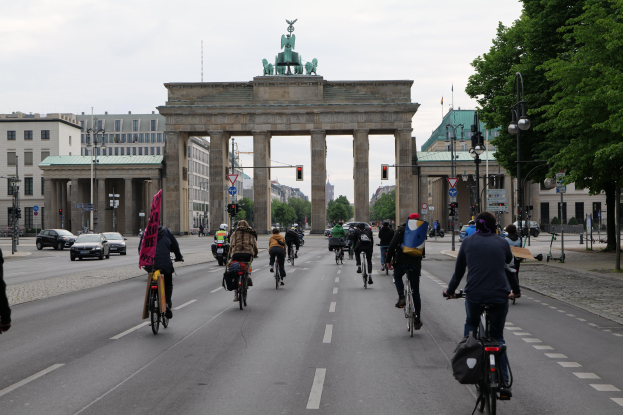 Eine Gruppe von Menschen, die Fahrräder vor dem Brandenburger Tor in Berlin, Deutschland, fahren, mit Bäumen, Laternenmasten und Gebäuden im Hintergrund unter einem bewölkten Himmel.
