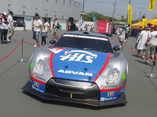 Ein Nissan GT-R Nismo GT3 Rennwagen auf einer Straße umgeben von Barrieren, Zuschauern, Zelten, Bannern und einem Gebäude unter einem bewölkten Himmel.