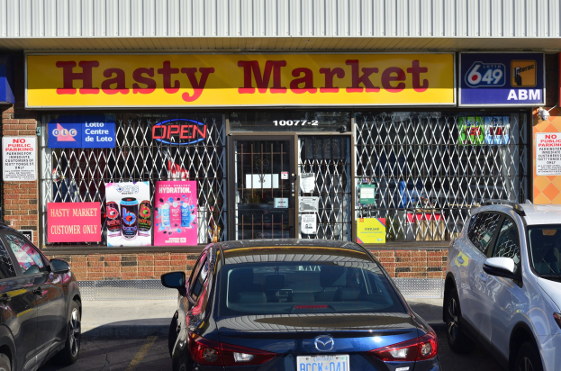 Ein geschäftiges Hasty Market-Geschäft in Toronto, Ontario, mit Autos auf der Straße davor, das Glaswände, -türen und Wandplakate zeigt.