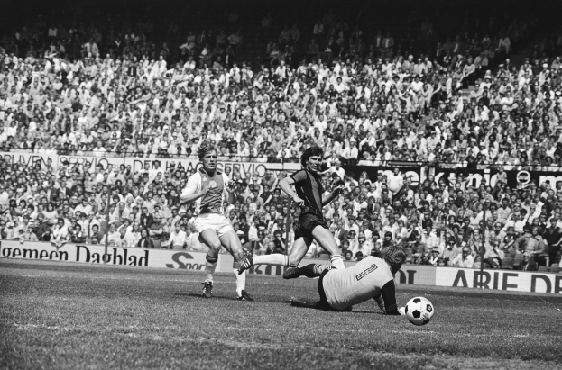 Schwarzes und weißes Foto von Männern beim Fußballspielen auf einem Feld mit Zuschauern im Hintergrund und einer 'Bundesliga 1978-79 Ajax 1-1 Hannover 96'-Fahne.