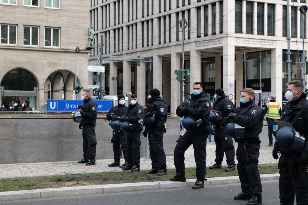 Polizisten in schwarzen Uniformen und Masken stehen vor einem Berliner Gebäude mit Glasfenstern und Säulen, einige halten Helme, mit Laternen, Verkehrsampeln, Schildern mit Text, einer Statue und grasbewachsenem Boden im Hintergrund.