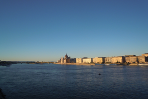 Ansicht des Donauufers in Budapest, Ungarn, mit Booten, Gebäuden, Bäumen, einer Brücke und Himmel im Hintergrund.