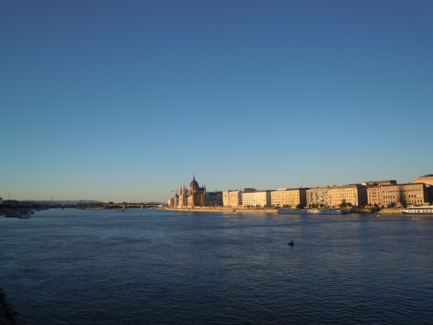 Ansicht des Donauufers in Budapest, Ungarn, mit Booten, Gebäuden, Bäumen, einer Brücke und Himmel im Hintergrund.