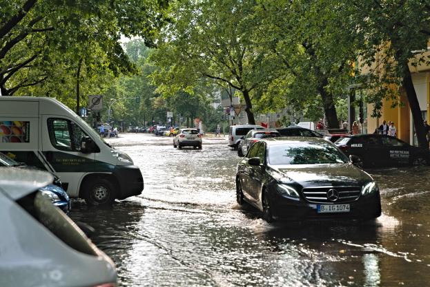 Überschwemmte Straße in Berlin, Deutschland mit Autos, die durch das Wasser fahren, Fußgängern auf dem Gehweg, Bäumen, Gebäuden und teilweise untergetauchten Schildern und Pfählen.