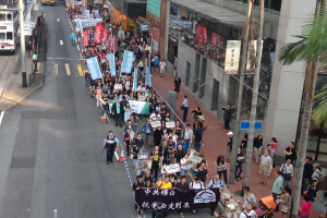 Eine große Gruppe von Menschen marschiert auf einer Stadtstraße und hält Schilder und Banner, während im Hintergrund Bäume, Gebäude und Fahrzeuge zu sehen sind.