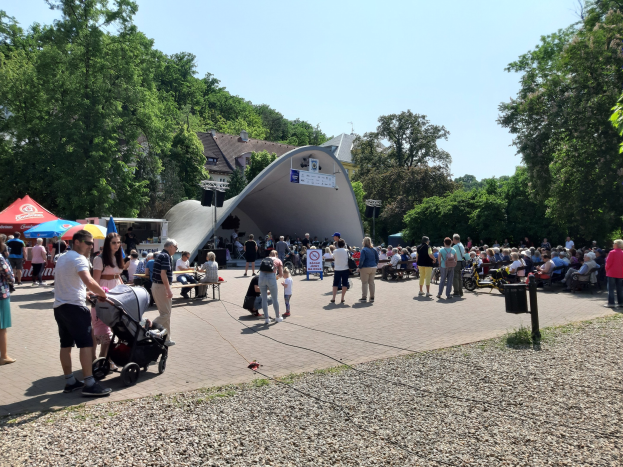 Eine Menschenmenge versammelt sich vor einem Gebäude bei einer Festival-Eröffnung, wobei einige auf Bänken sitzen und andere stehen, einschließlich einer Person mit einem Kinderwagen im Vordergrund. Bäume, Gebäude und ein klarer blauer Himmel füllen den Hintergrund, während Zelte die linke Seite säumen und ein Müllcontainer auf der rechten Seite zu sehen ist.