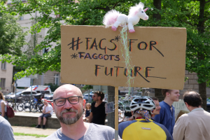 Ein Mann mit Brille hält ein Schild mit der Aufschrift "Fakten für Schwuchteln Zukunft" mit einem Spielzeug darauf, umgeben von Menschen, Fahrrädern, Fahrzeugen, Bäumen und Gebäuden.