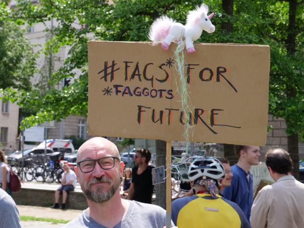 Ein Mann mit Brille hält ein Schild mit der Aufschrift "Fakten für Schwuchteln Zukunft" mit einem Spielzeug darauf, umgeben von Menschen, Fahrrädern, Fahrzeugen, Bäumen und Gebäuden.