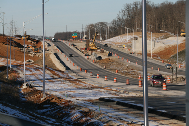 Autobahn mit Baumaschinen am Straßenrand, darunter Fahrzeuge, Verkehrskegel, Pfosten, Lampen, Schilder und andere Gegenstände, vor schneebedeckter Landschaft, Bäumen und einem klaren Himmel.
