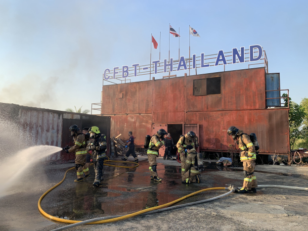 Feuerwehrleute in Helmen sprühen Wasser auf ein Gebäude mit Text, während Fahnen, Bäume und ein klarer blauer Himmel im Hintergrund zu sehen sind.