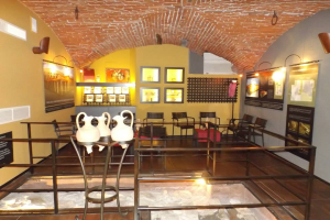 Ein Museumsraum voller Tische, Stühle, einem Geländer, Vasen, Texttafeln, Wandrahmen, Lampen und verschiedenen Ausstellungsgegenständen.