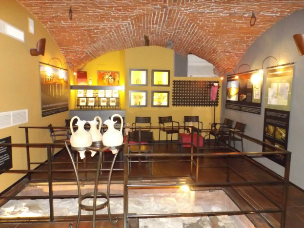 Ein Museumsraum voller Tische, Stühle, einem Geländer, Vasen, Texttafeln, Wandrahmen, Lampen und verschiedenen Ausstellungsgegenständen.