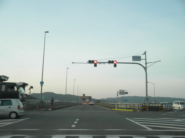 Ein Auto fährt auf einer Autobahn an einer Kreuzung mit einer Ampel vorbei, umgeben von Fahrzeugen, einem Fußgänger auf dem Gehweg, Geländern, Laternen, Verkehrszeichen, Straßenschildern, Gebäuden, Hügeln und einem klaren blauen Himmel.