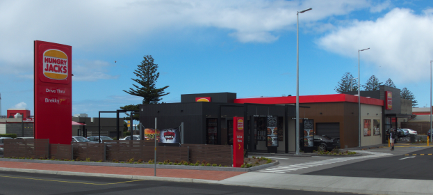 Eine belebte städtische Straßenkreuzung mit Gebäuden, Bäumen, Straßenmöbeln, Fahrzeugen, Fußgängern und einem Hungry Jacks Restaurant im Vordergrund.