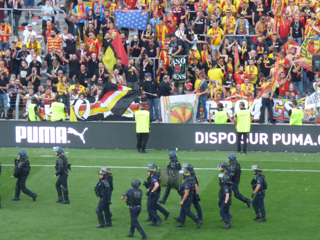 Polizisten in Helmen und taktischer Ausrüstung überqueren ein Fußballfeld, bewaffnet, mit Zuschauern, die Fahnen und Banner halten.