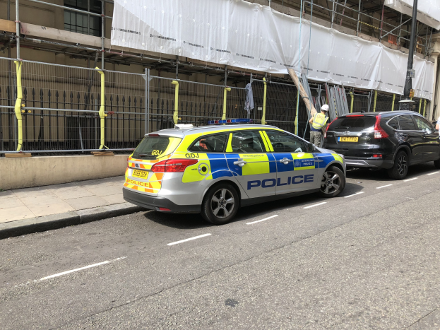 Polizeiauto auf einer Straße neben einem Gebäude mit zwei Helikopterpiloten und einem weißen Tuch, das wahrscheinlich an einem Tatort in London drapiert ist.