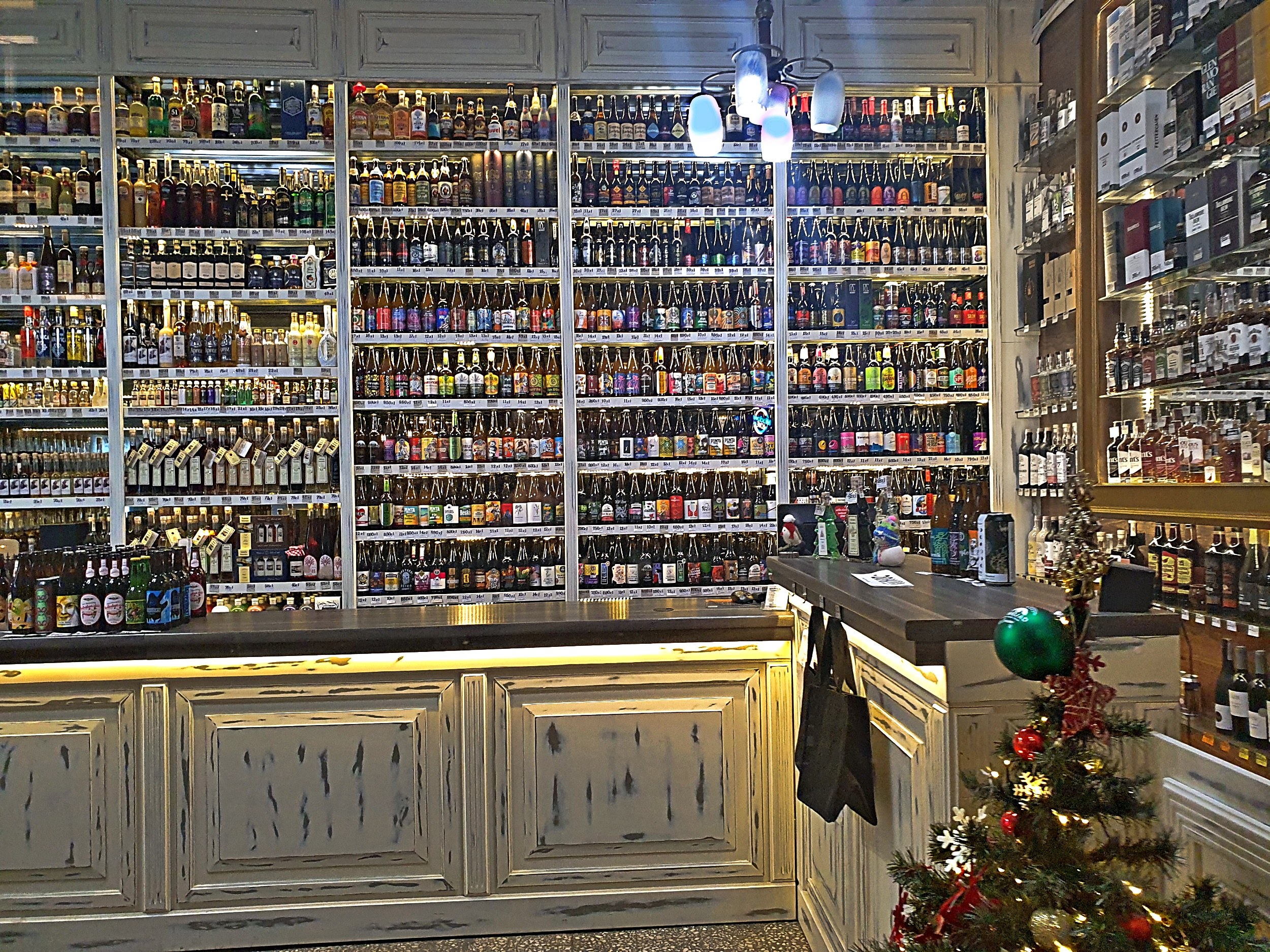 Ein Geschäft mit Regalen voller Spirituosenflaschen, einem geschmückten Weihnachtsbaum, einer Kommode mit Schubladen und von der Decke hängenden Lichtern.