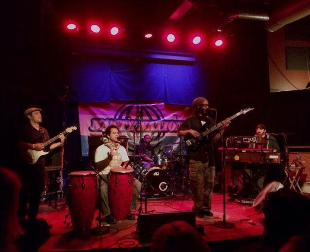 Eine Gruppe von Musikern, die auf der Bühne mit Instrumenten, Mikrofonen und Ständern spielt, beleuchtet von Scheinwerfern, mit einem Publikum im Vordergrund.