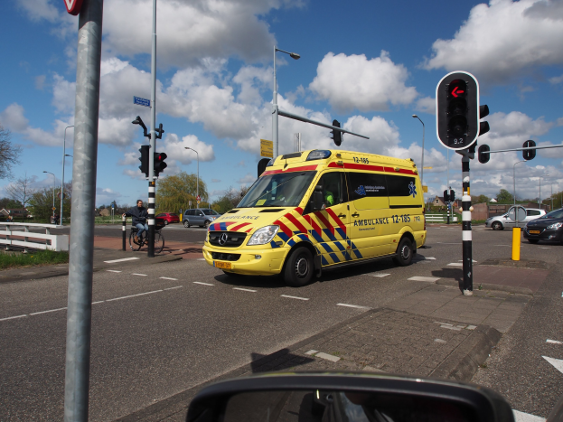 Ambulanz fährt auf einer Straße neben einer Ampel, mit einem Radfahrer im Vordergrund und Gebäuden, Bäumen, Laternen und einem bewölkten Himmel im Hintergrund; ein Spiegel ist unten zu sehen.