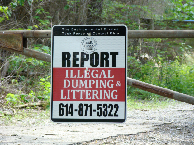 Ein Schild am Straßenrand trägt die Aufschrift "Meldet Illegalen Müll und Littering", mit Bäumen und Pflanzen im Hintergrund und einer Tafel mit Text im Vordergrund.