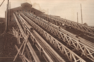 Ein altes Schwarz-Weiß-Foto einer hölzernen Achterbahn, die eine Schiene hinunterfährt, mit einem Gebäude mit Fenstern, Strommasten mit Drähten und einem Himmel im Hintergrund, mit Text am unteren Bildrand.