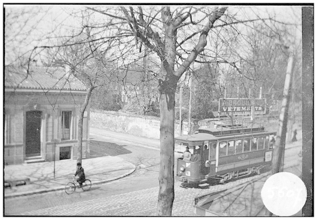 Schwarzes und weißes Foto einer Straßenbahn auf einer Stadtstraße mit einem Fahrradfahrer vorne, Passagiere im Inneren sichtbar und Bäume und ein Gebäude im Hintergrund.