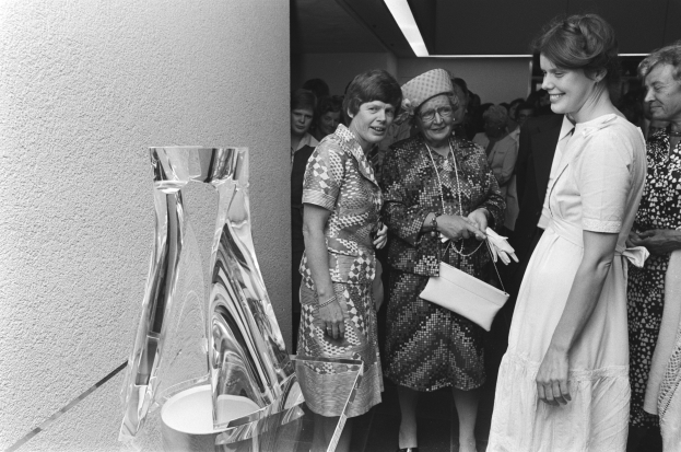 Eine Gruppe von Frauen steht zusammen vor einer Skulptur, mit einer Wand links und Lichtern oben, dargestellt in Schwarz-Weiß.