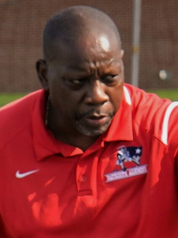 Ein Mann in einem roten T-Shirt steht auf einem Feld und hat die Hände in die Hüften gestemmt, er sieht ernst aus mit einem unscharfen Hintergrund.