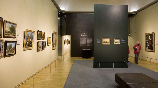 Großer Raum mit zahlreichen Gemälden an den Wänden, zwei Personen stehen, eine Couch, ein Tisch mit Gegenständen, eine Tafel mit der Aufschrift "Karel du Jardin Ausstellung in der Nationalgalerie der Künste" und Deckenbeleuchtung.