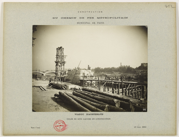 Schwarz-weiß-Foto einer Baustelle mit Holzstämmen, einer Brücke, einem Turm, Bäumen und einem Himmel im Hintergrund, mit einem Stempel auf dem Foto.