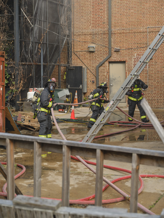 Feuerwehrleute mit Helmen arbeiten daran, ein Gebäude Feuer zu löschen, umgeben von Ausrüstung, einem Zaun und Hintergrundelementen einschließlich eines Baums und des Himmels.