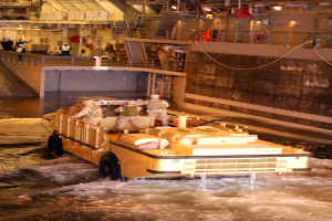 Amphibisches Fahrzeug mit Helmtr├Ągern durch Wasser in der N├Ąhe einer Wand, mit Menschen, Gel├Ąndern, Lichtern und Rohren im Hintergrund.