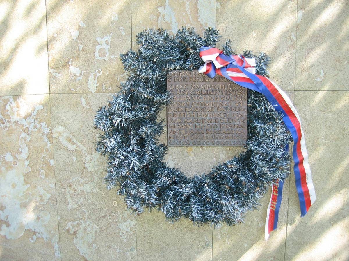 Ein Kränzchen mit einem Kranz und einer Inschrift hängt an einer Gebäudewand, geschmückt mit einer rot-weiß-blauen Schleife.